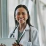 Female doctor holding digital tablet, smiling at camera beside window at corridor in hospital